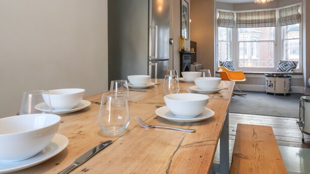 Ein Essbereich mit einem Holztisch, gedeckt für vier Personen, einschließlich Schüsseln, Tellern, Gläsern und Besteck. Im Hintergrund sind ein moderner Kühlschrank und eine Kristallleuchter zu sehen.