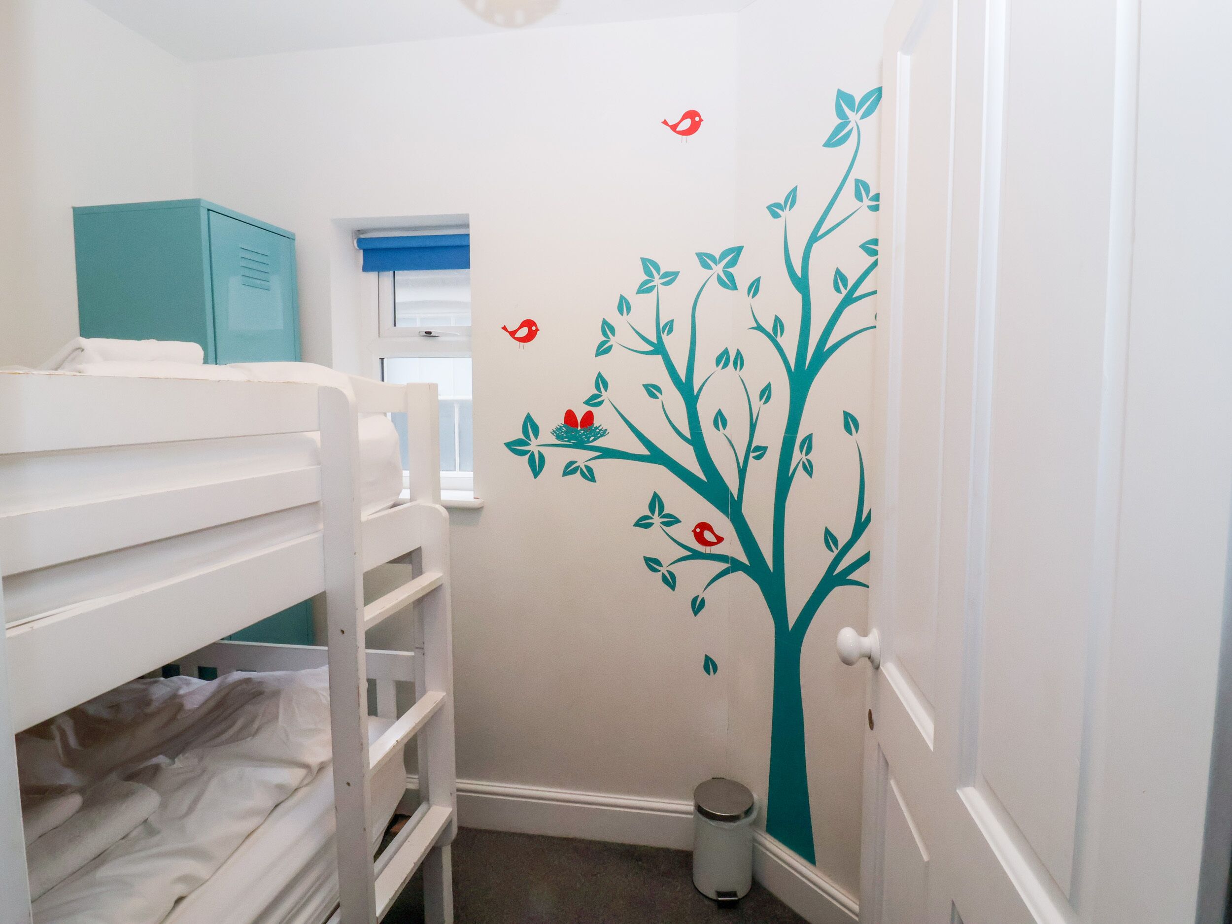 A small, narrow room with bunk beds, a window with blue blinds, a wall with a tree decal, and a door.