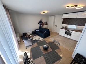 A modern living room with a sofa, coffee table, and dining table. There is a kitchen area with cabinets and appliances. - Appartement 6 personnes, 5mn Disneyland Paris, Val d'Europe + Captain America ++ (Chessy)
