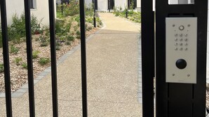 A gated entrance to a modern building with a keypad lock, a gravel pathway, and a view of multi-story residential units with balconies. - Appartement 6 personnes, 5mn Disneyland Paris, Val d'Europe + Captain America ++ (Chessy)