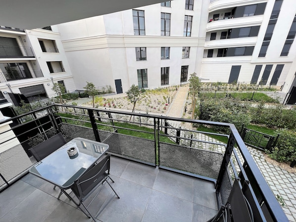 A balcony with a glass table and chairs, overlooking a courtyard with plants and a pathway. - Appartement 6 personnes, 5mn Disneyland Paris, Val d'Europe + Captain America ++ (Chessy)