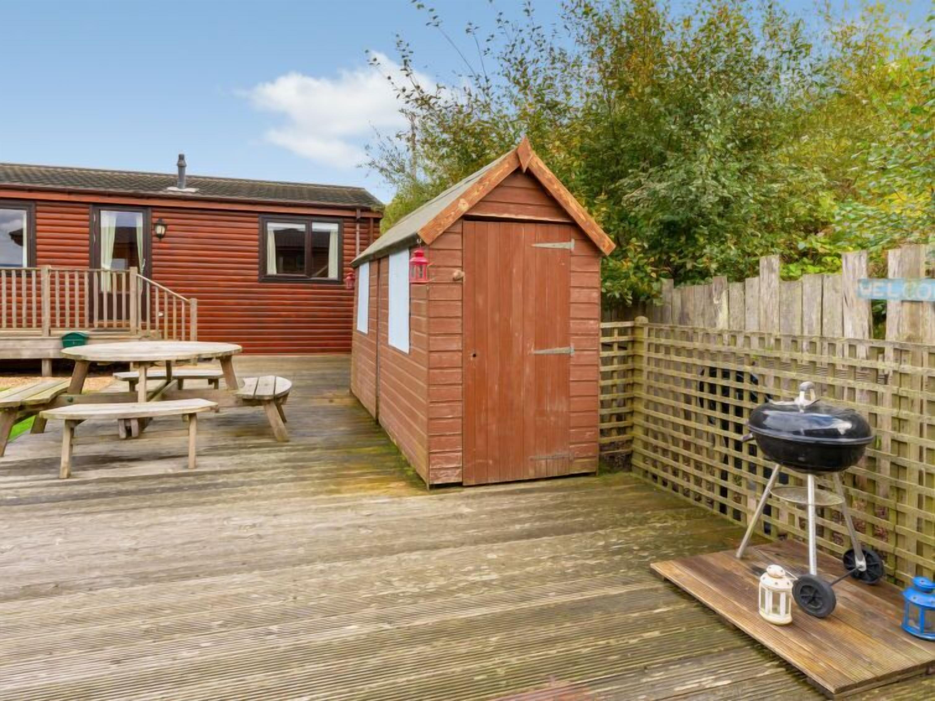 Une terrasse en bois avec des tables de pique-nique, un barbecue et un petit abri de jardin.