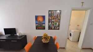 Uma sala de jantar com mesa preta e cadeiras laranjas, um armário preto com televisão e um cartaz na parede.