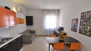 Cozinha moderna com armários cinza e preto, sofá cinza e uma mesa de jantar com cadeiras laranja. Há uma TV em um suporte preto e uma janela com cortinas brancas.
