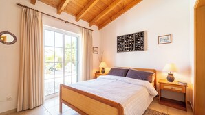 Un dormitorio con cama de madera, dos mesitas de noche con lámparas, un ventanal con cortinas y una estantería para vinos en la pared.