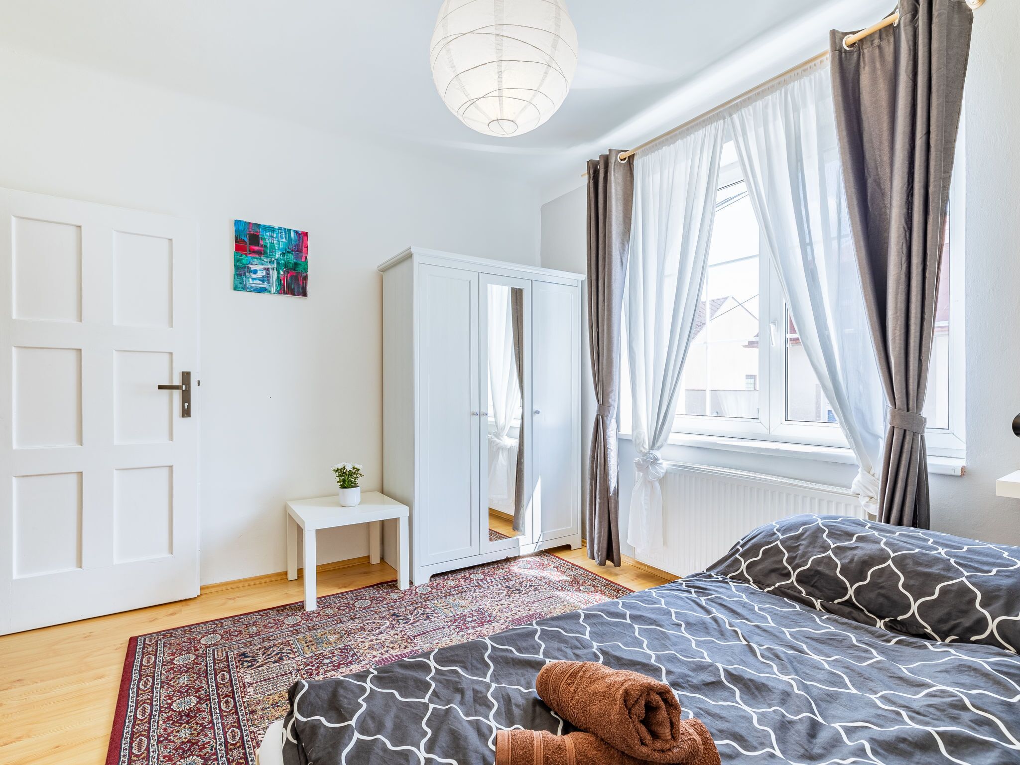 A bedroom with a bed, a small bedside table, a mirror, a wardrobe, and a window with curtains.