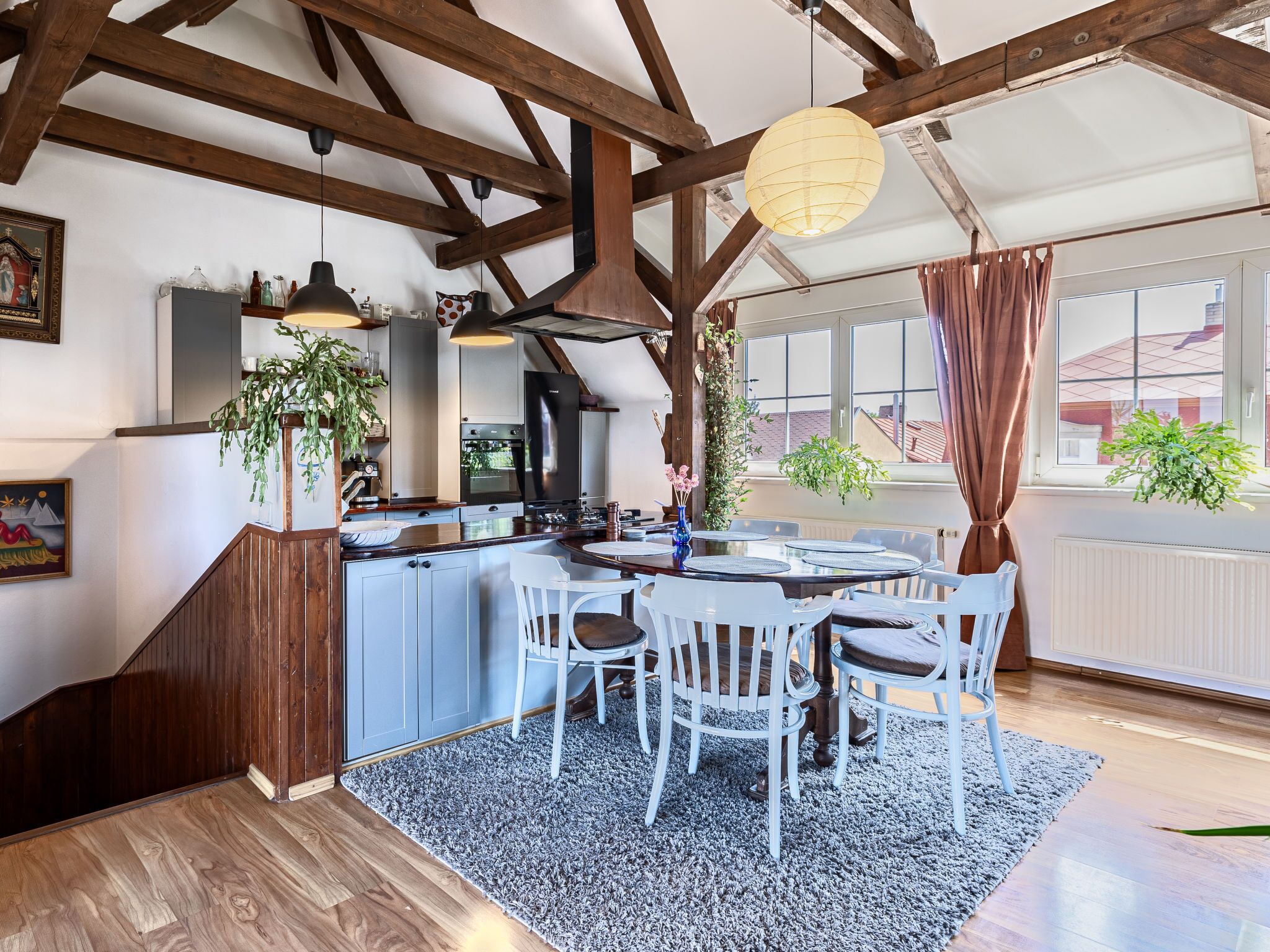 A kitchen with wooden beams, a dining area with white chairs, and a large window with curtains.