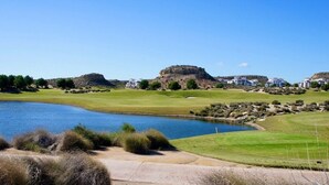 Camp de golf amb un estany, un turó gran i diversos edificis blancs al fons.