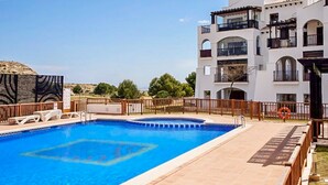 Una piscina amb un solàrium de fusta i gandules, envoltada per una tanca i un edifici de diversos pisos amb balcons.