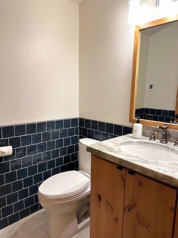A bathroom with a white toilet, dark blue tiled walls, a wooden vanity with a sink, and a mirror above the sink.