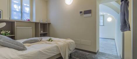 A bedroom with a bed, a radiator, a window, and a door leading to another room.