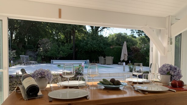 Un espace de restauration en plein air avec une table en bois dressée pour un repas, une bouteille de vin et un saladier de fruits.