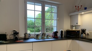 Une cuisine moderne avec une grande fenêtre, divers appareils électroménagers et un saladier rempli d’oranges posé sur le plan de travail.
