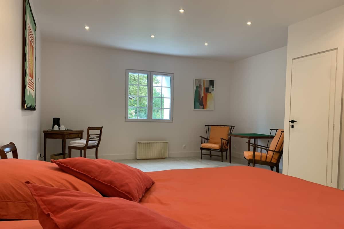 Une chambre à coucher avec un grand lit, deux chaises, une petite table, un radiateur et une fenêtre donnant sur un paysage verdoyant.