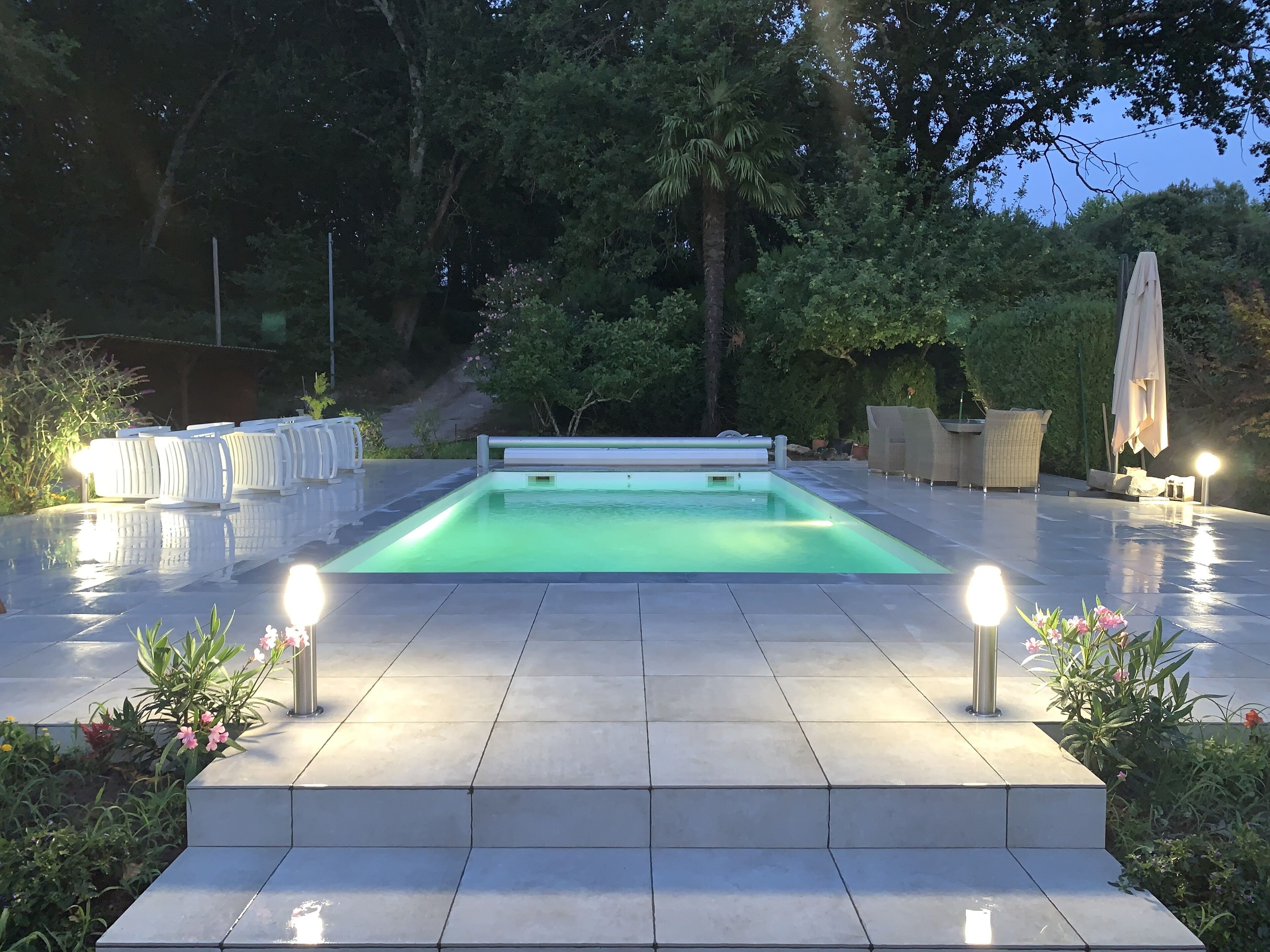 A swimming pool with a tiled deck, surrounded by greenery and outdoor furniture.