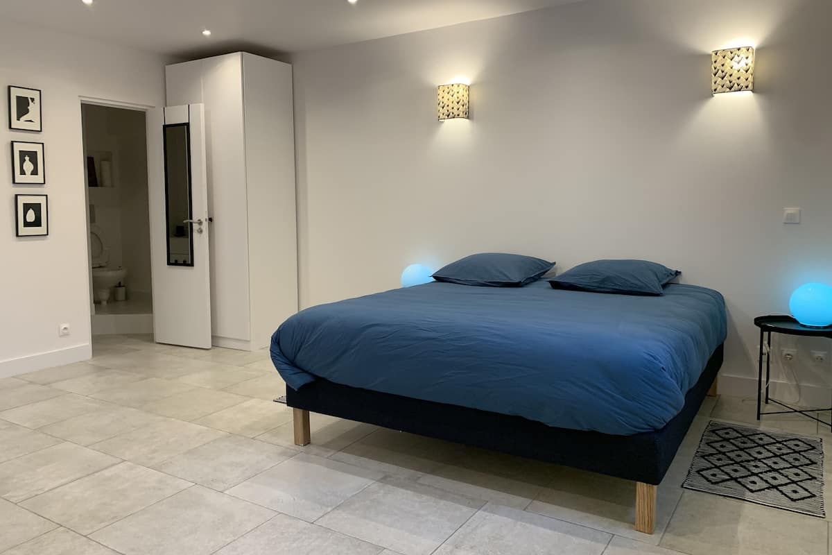Une chambre à coucher comprenant un lit, une table de chevet, une lampe fixée au mur et une porte donnant sur une salle de bain.