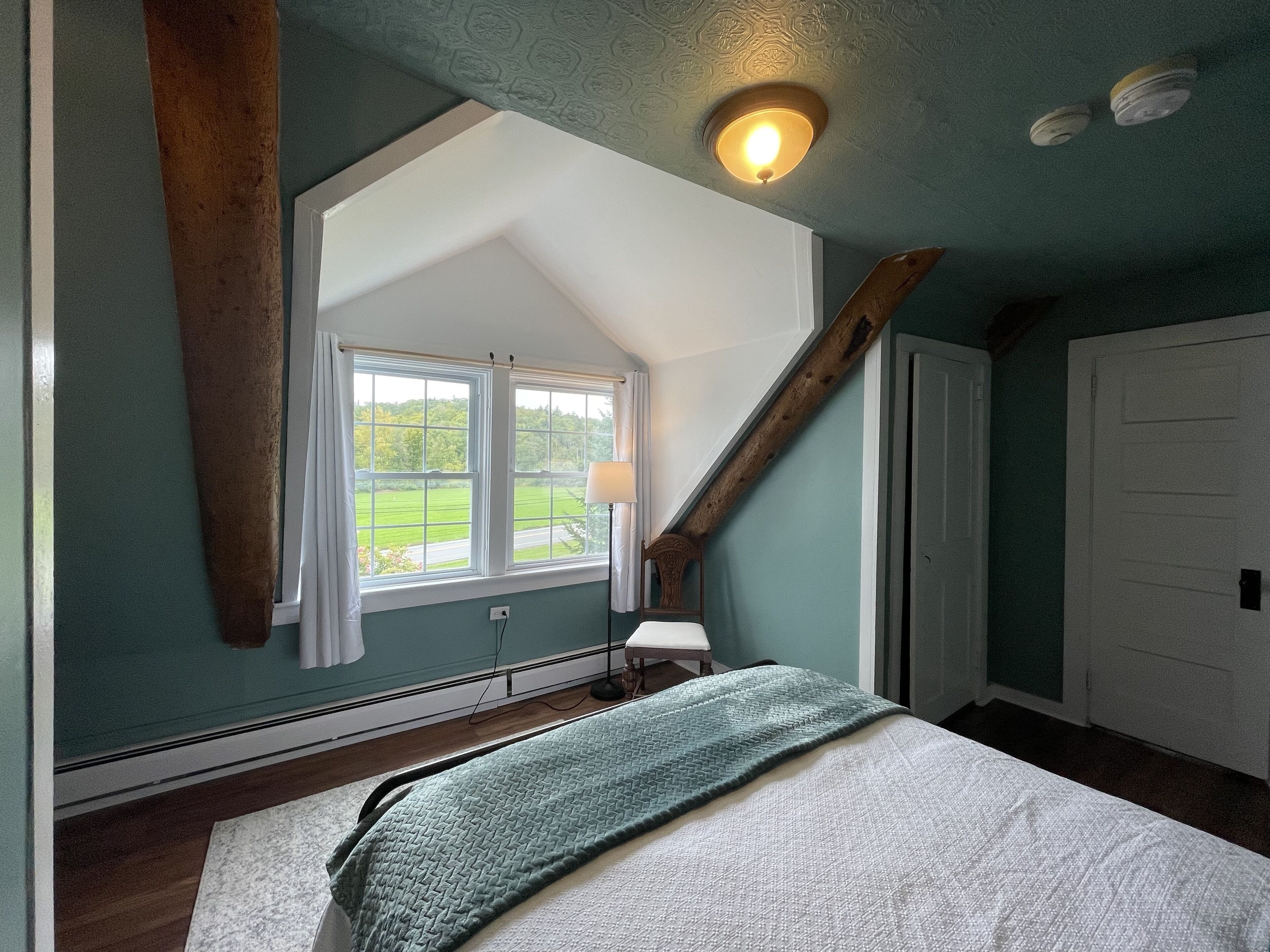A bedroom with a sloped ceiling, a window with curtains, a bedside lamp, and a bed with a green blanket.