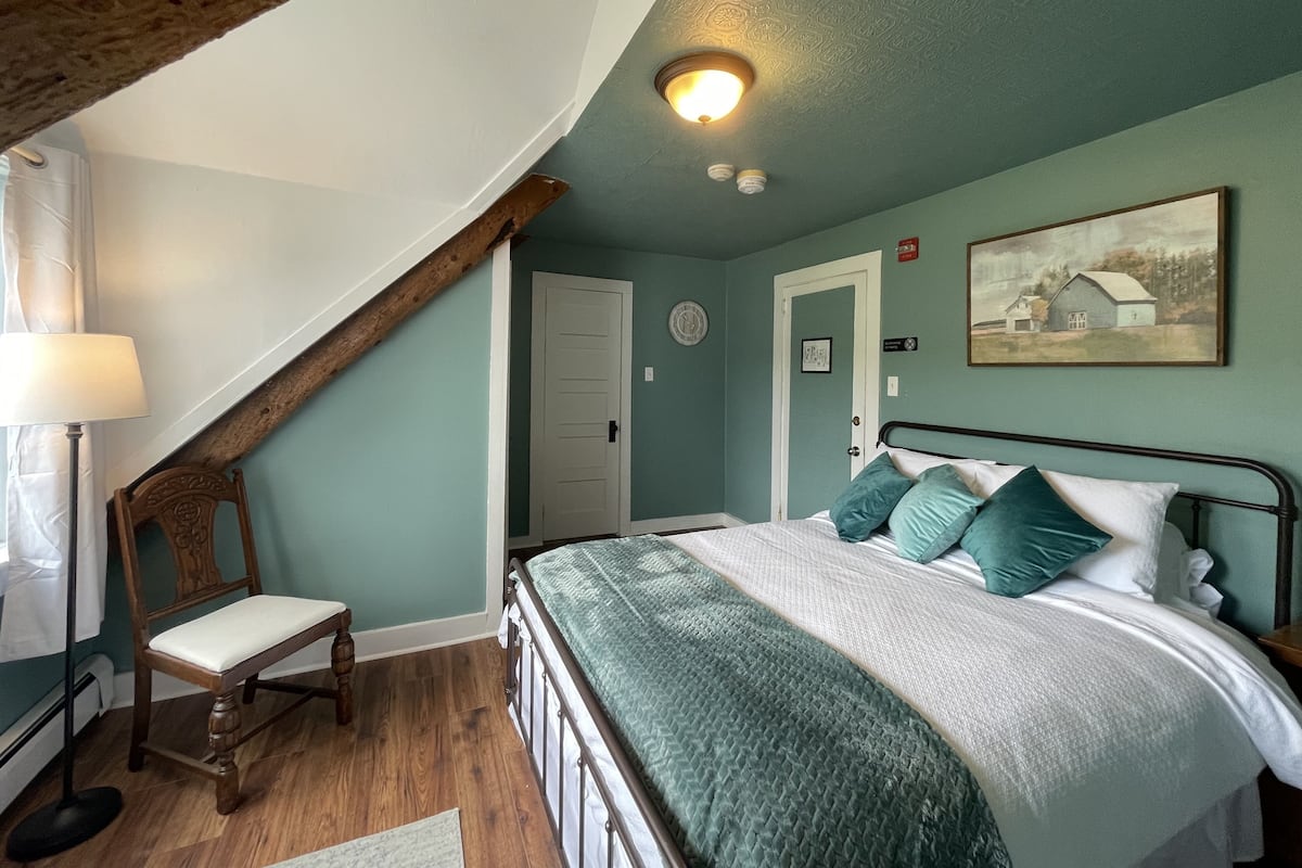 A bedroom with a sloped ceiling, a bed with green and white bedding, a wooden chair, and a painting on the wall.