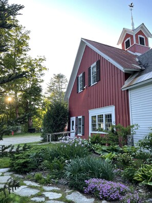 A red barn with a white door and shutters, surrounded by a garden with various plants and flowers. - Stone Hearth Inn and Eatery (Chester)
