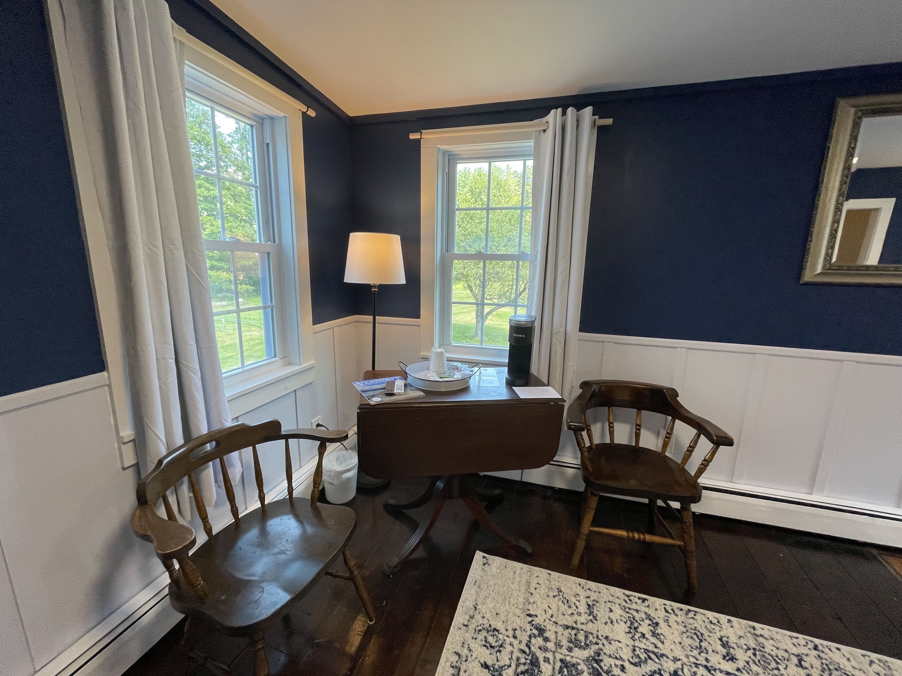 A room with two wooden chairs, a small table, a lamp, and a window with a view of greenery.