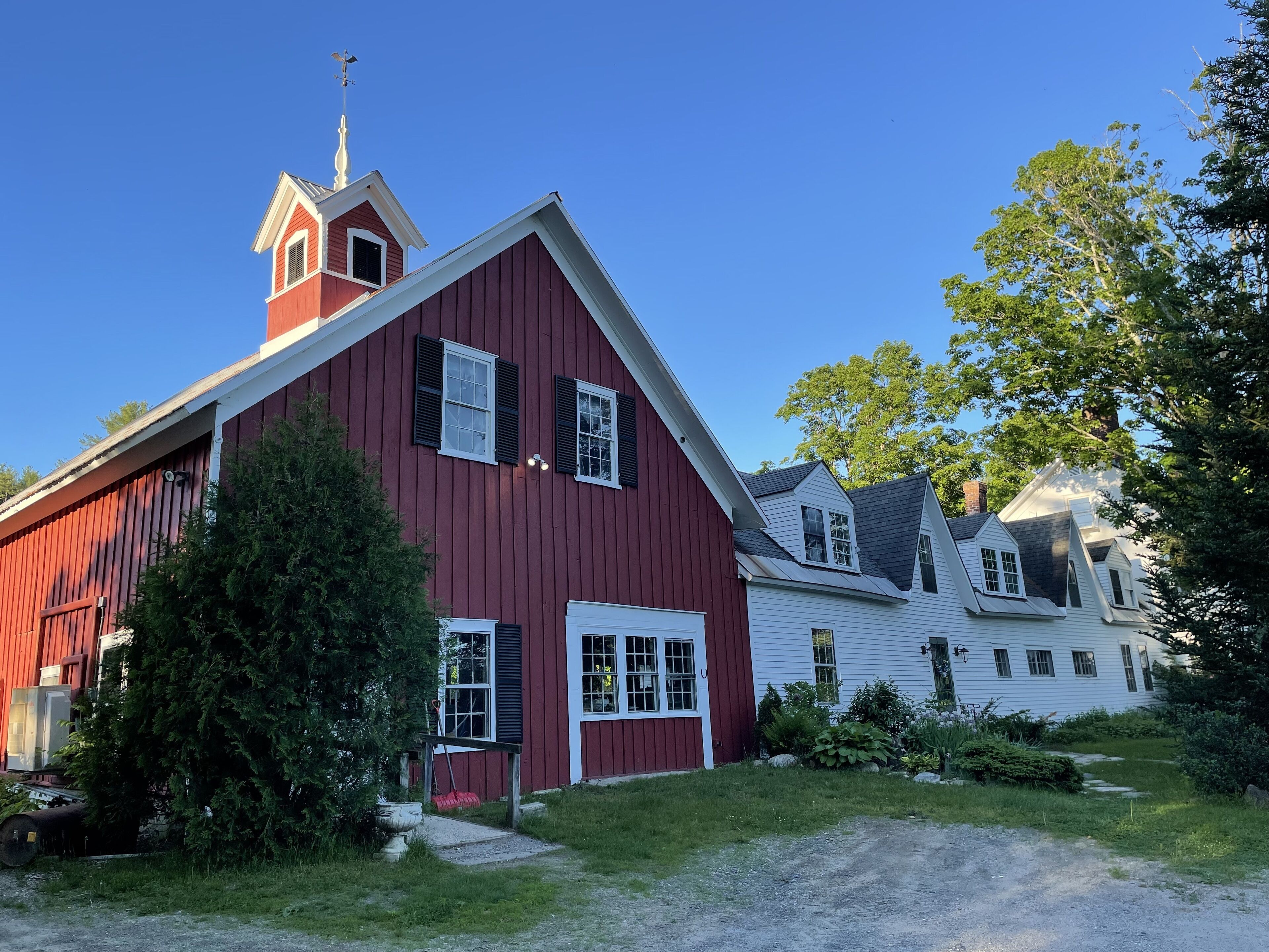 A red barn with a white door and a small attached structure, surrounded by greenery and a gravel path.
