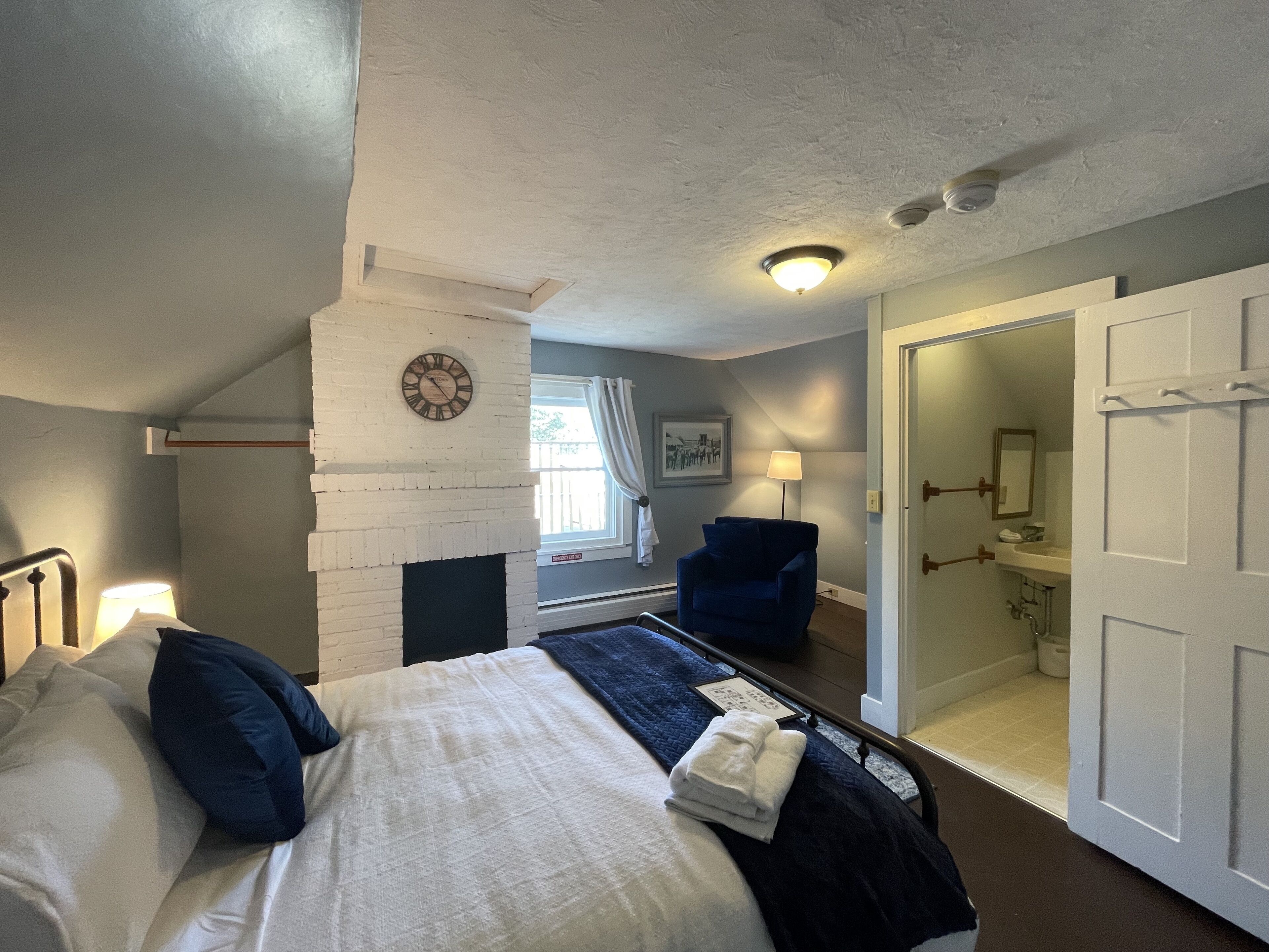 A bedroom with a bed, a chair, a clock, a window, and a door leading to a bathroom.