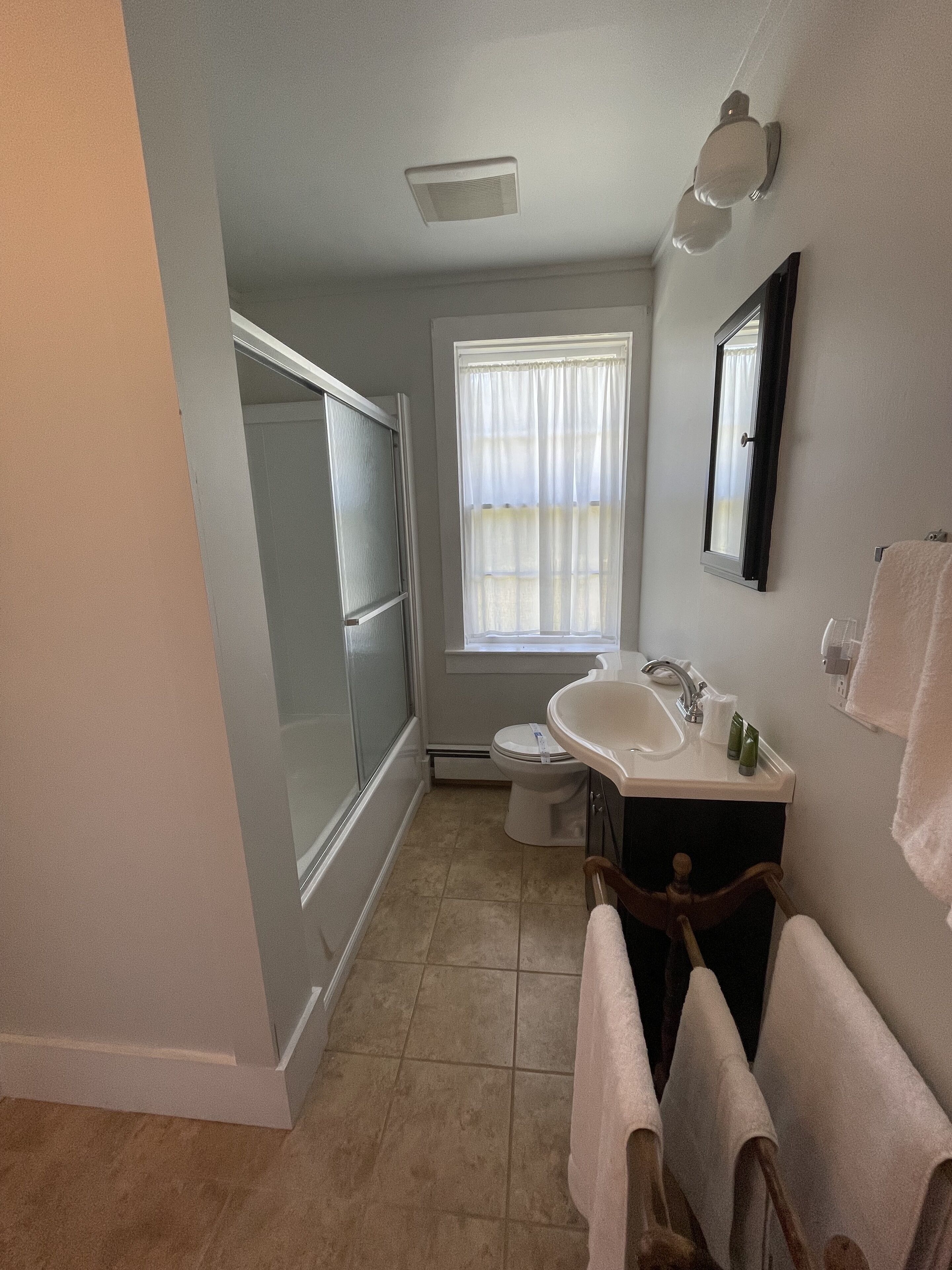 Baño con ducha, inodoro, lavabo y ventana con cortinas.