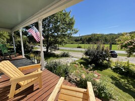 Una terraza de madera con dos sillas Adirondack, una bandera estadounidense y vistas de un paisaje verde con una carretera y colinas.