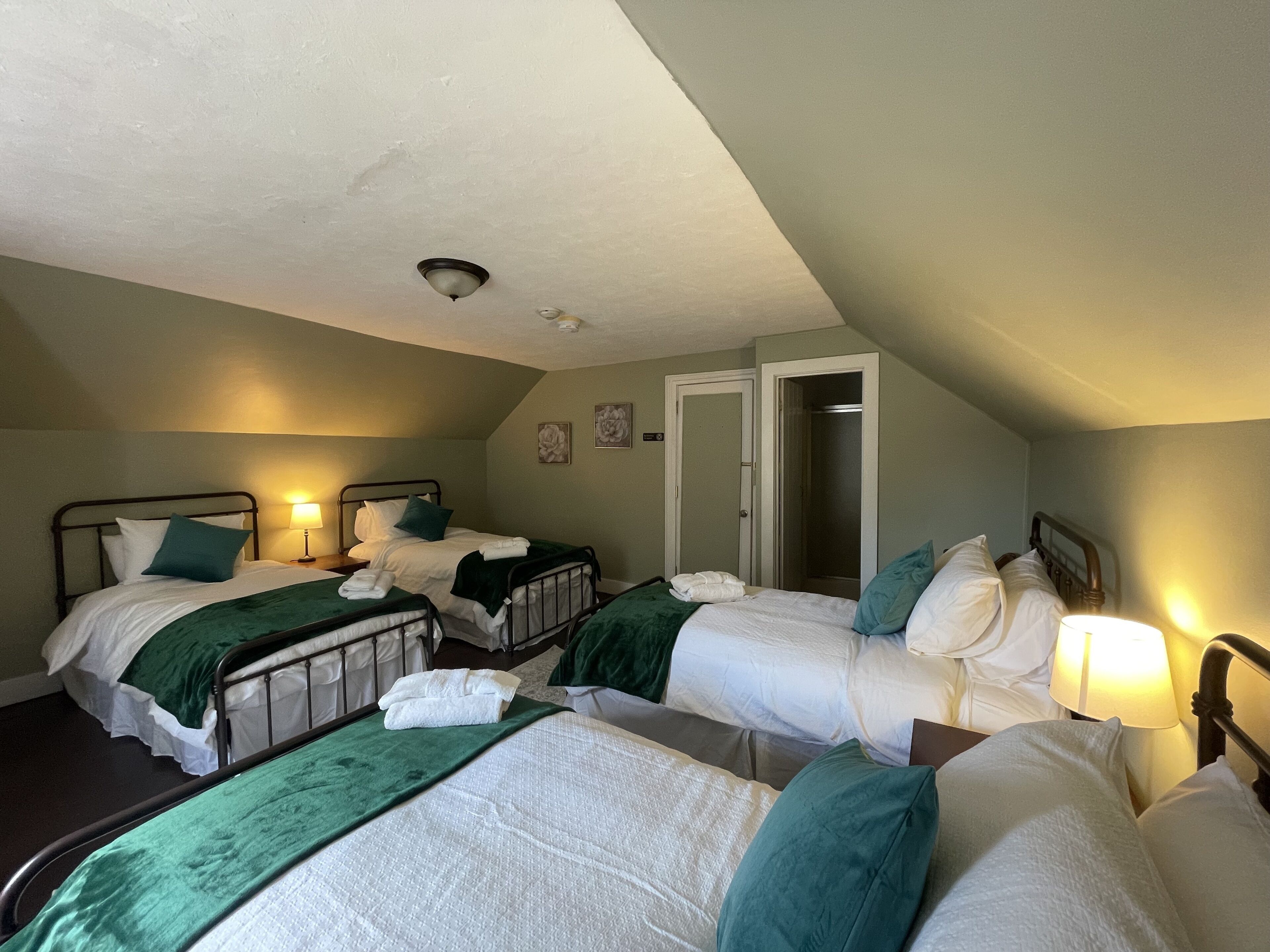 Habitación de hotel con tres camas dispuestas en fila, cada una con ropa de cama blanca y almohadones verdes. Hay una puerta que da a un armario y una ventana pequeña en la pared de la izquierda.