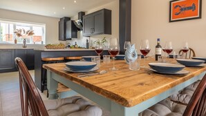 A modern kitchen with dark cabinets, a wooden dining table set for six, and a large window providing natural light. - Sandpiper House (4), Ingoldisthorpe, Norfolk (Ingoldisthorpe)