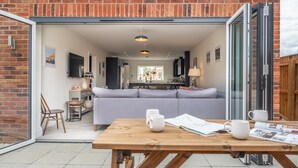 A modern kitchen with a grey sofa, a wooden table with mugs and papers, and a brick exterior wall. - Sandpiper House (4), Ingoldisthorpe, Norfolk (Ingoldisthorpe)