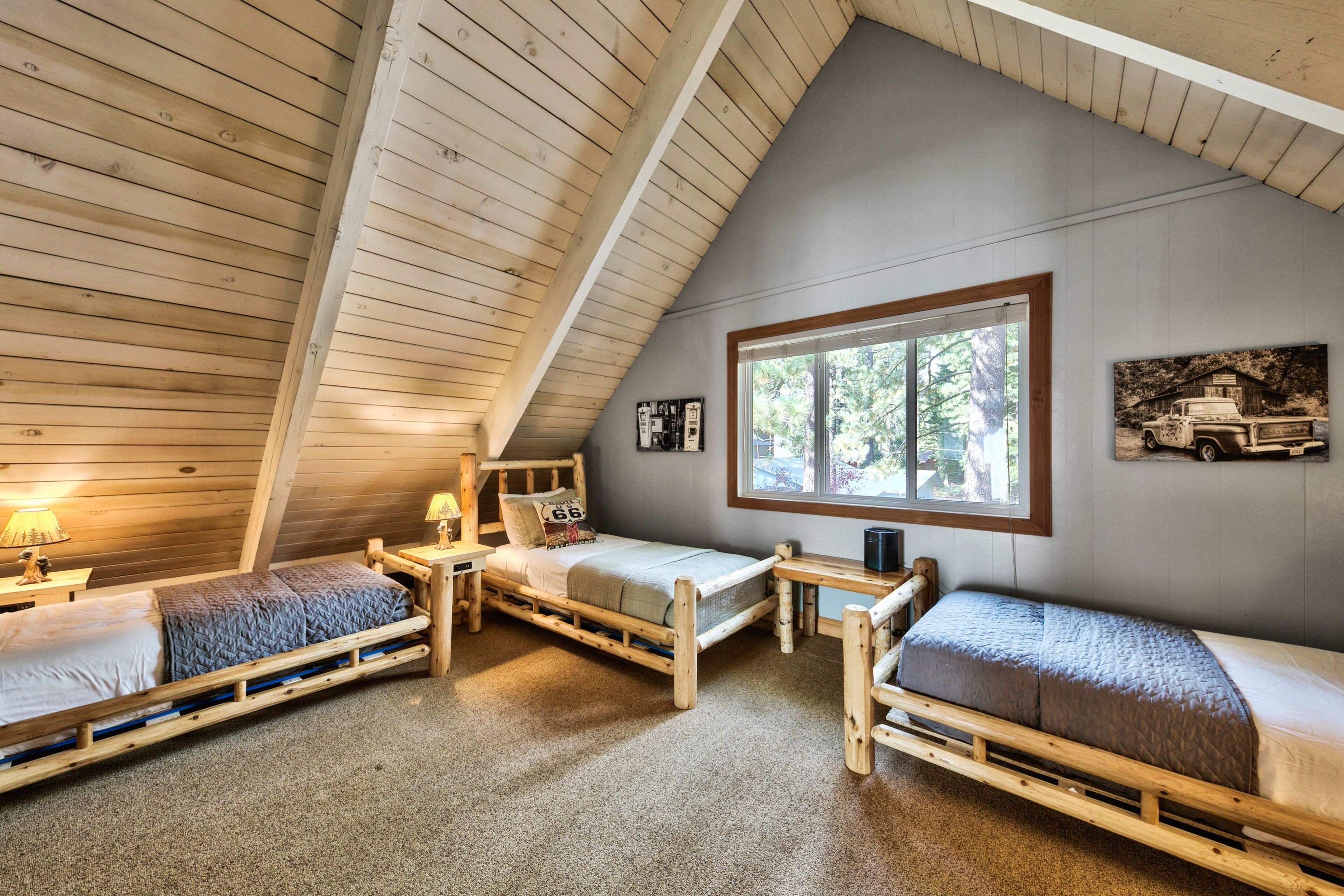 Un dormitorio abrigado en el ático con tres camas de madera, un ventanal grande y una decoración rústica que incluye un cuadro de un camión.