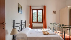Een tweepersoonsbed met een witte spreideken, een nachtkastje met een lamp, een raam met oranje gordijnen en een klein schilderij aan de muur.