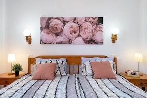 Una cama doble con ropa de cama estampada, una obra de arte en la pared con flores rosas, lámparas de noche y una pequeña planta en maceta.