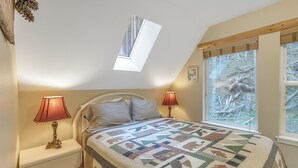 Un dormitorio con techo inclinado, tragaluz, cama con colcha, dos lámparas de noche y ventana con vista al bosque.