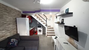 Een compacte leefruimte met een bank, een flatscreen televisie, een wasmachine en een trap naar een keuken.