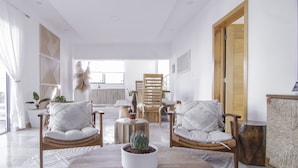 A spacious living room with a large window, a wooden dining table, two cushioned chairs, a coffee table with a plant, and a wooden cabinet. - New Beachfront Penthouse by RedAwning (Las Terrenas)