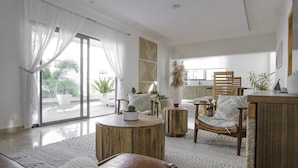 A spacious living area with a large sliding glass door, a wooden coffee table, a wicker chair, and a small side table. - New Beachfront Penthouse by RedAwning (Las Terrenas)