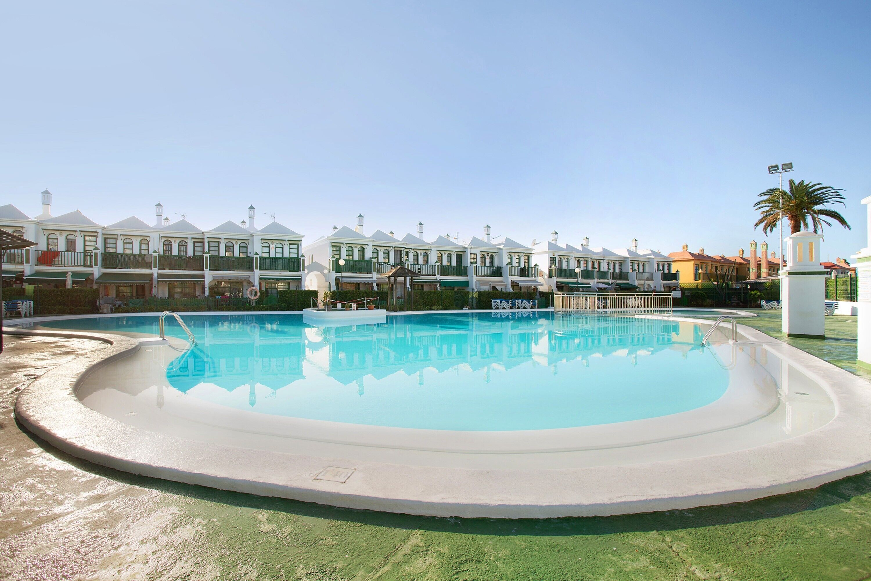 Une piscine entourée de bâtiments aux façades uniformes, dotés de balcons, avec un palmier et un ciel dégagé.