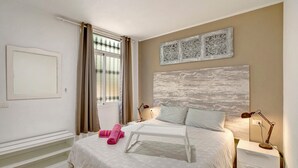 Un dormitorio con cama, mesita de noche con lámpara, ventana con cortinas y una obra de arte colgada en la pared.