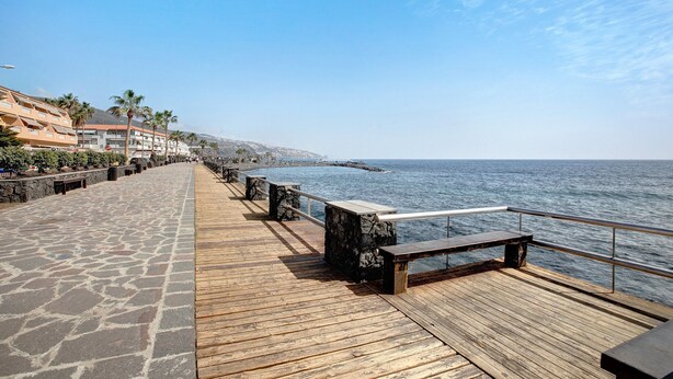 Una pasarela de madera que conduce al mar, con adoquines de piedra, bancos y palmeras.