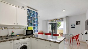 Cocina compacta con lavadora, microondas y una pequeña zona de comedor con sillas rojas.
