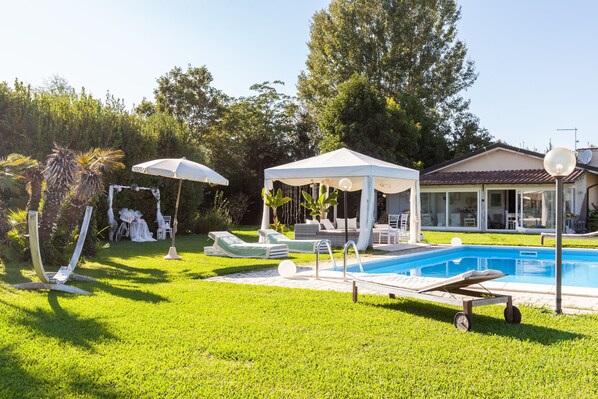 A pool area with lounge chairs, a gazebo, and a house with a tiled roof.