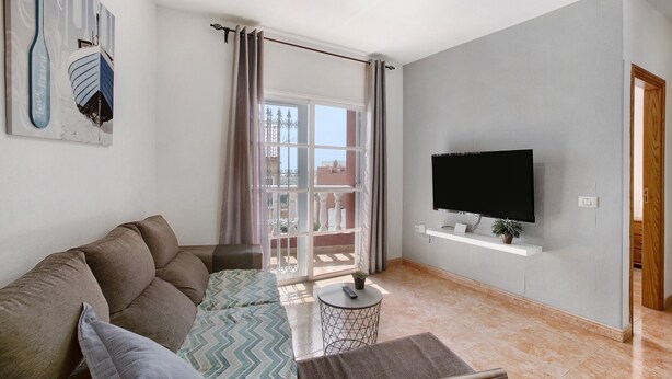 Un salon avec un canapé, un téléviseur fixé au mur, une petite table ronde et une fenêtre avec des rideaux.