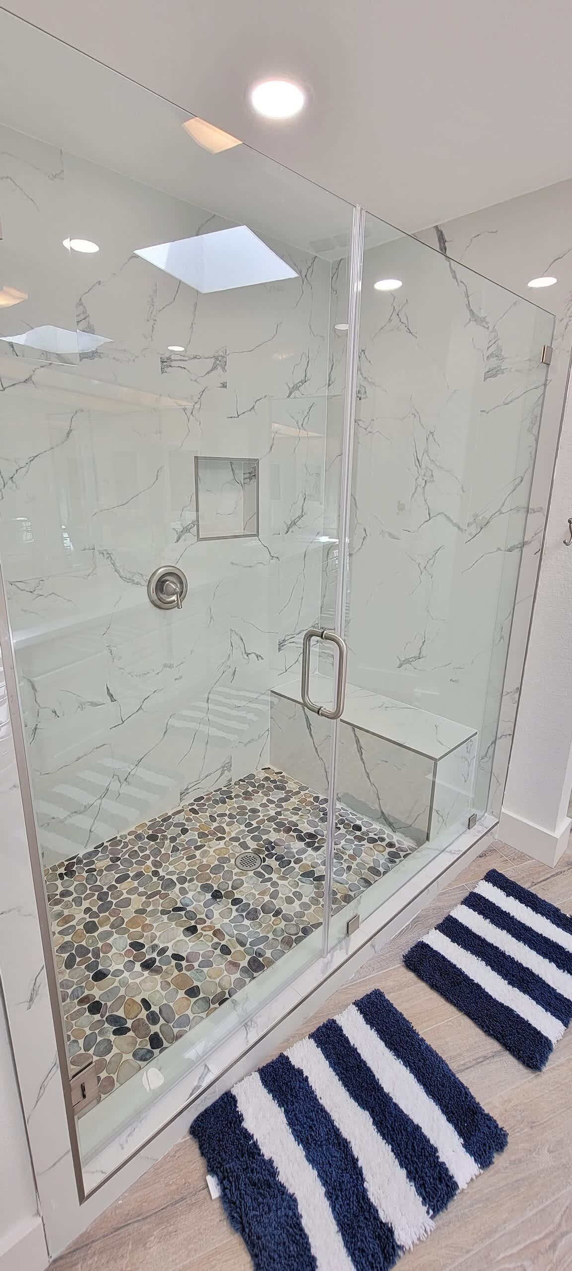 A glass-enclosed shower with a marble wall, a small shelf, and a tiled floor.