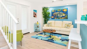 Una sala de estar con una escalera blanca, un sofá color beige, una mesa de centro y un cuadro abstracto y colorido en la pared.