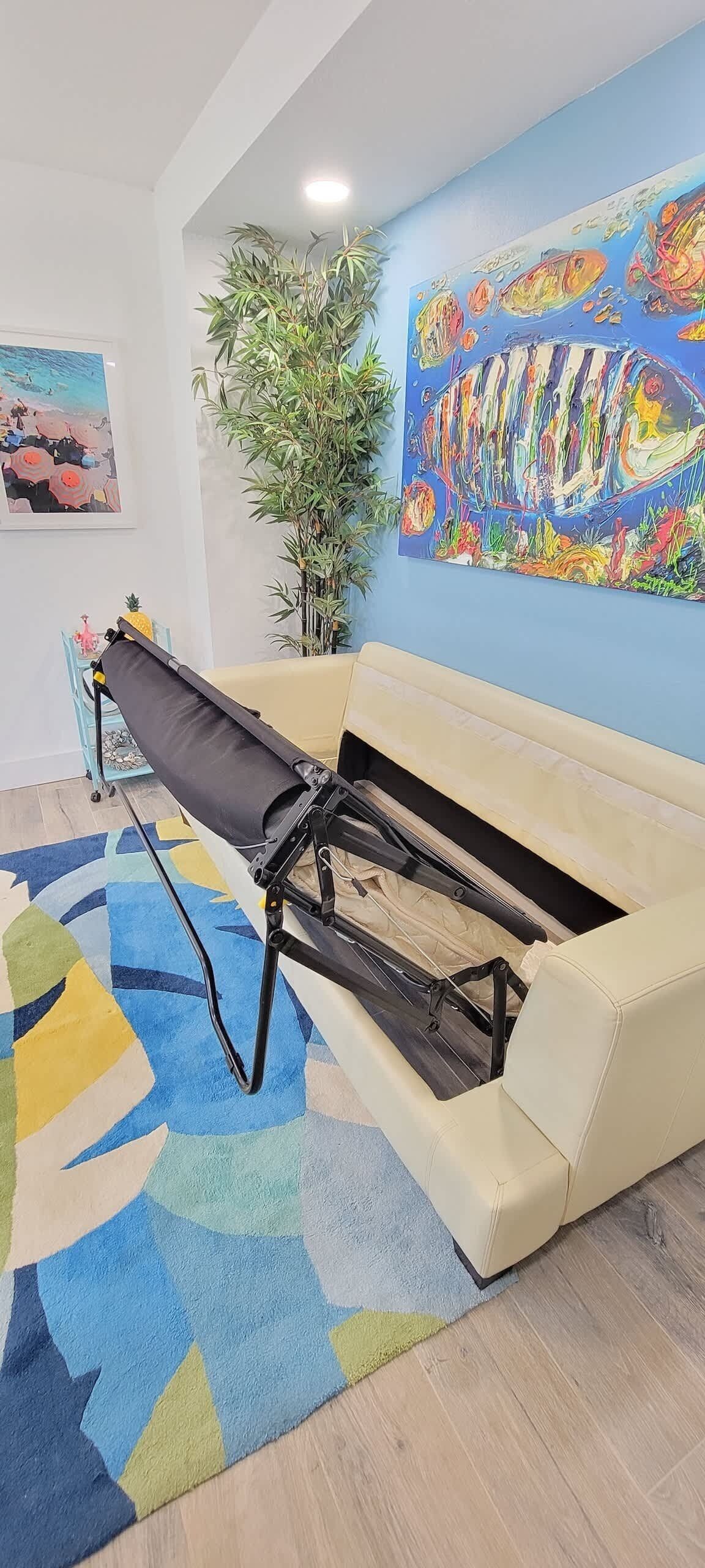 Ein beiges Sofa mit einem schwarzen Massagetisch davor, ein farbenfrohes abstraktes Gemälde an der Wand und eine große, grüne Pflanze.