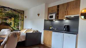 A compact, modern room with a bed, a dining area, a kitchenette, and a small garden view. - '12 MN Megève Studio Comfort' with Mountain View (Flumet)