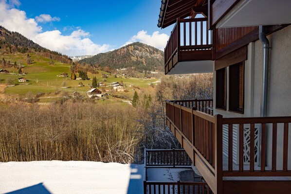 A mountain view from a balcony with a wooden railing, a snow-covered ground, and a clear sky.