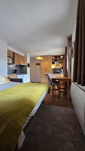 A compact room with a bed, a desk, a chair, a kitchenette, and a bookshelf.